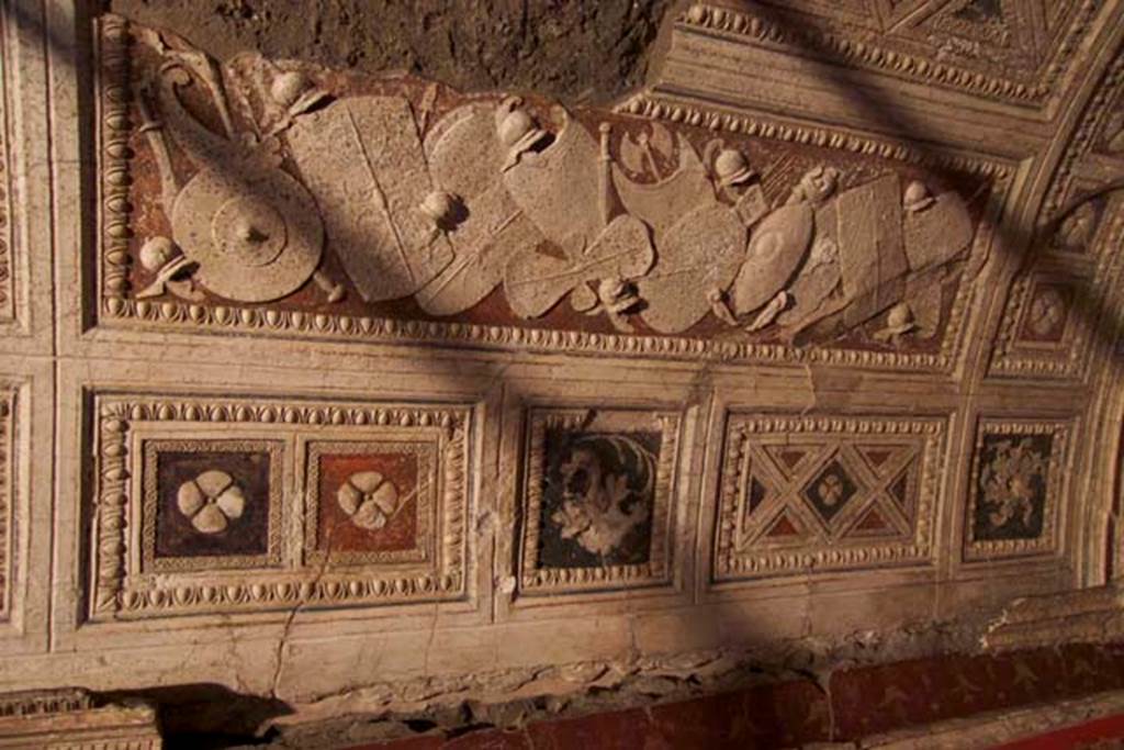 Villa dei Papiri, Herculaneum. July 2010. Room (I) on lower terrace, east side of vaulted stucco ceiling with frieze showing a pile of weapons. Photo courtesy of Michael Binns.
The weapons are represented with extreme accuracy in white stucco on a red background.
The weapons are predominantly north European, but the are also Hellenistic, eastern and barbarian weapons. Defensive weapons include a breastplate with corset, two greaves, nine helmets and fourteen shields of different shapes and sizes including a rare figure of eight shield with two concavities. The shields have incised decorations realized with a stylus.
Offensive weapons include a two-edged axe, a gorytos (quiver) with a sheaf of arrows, two swords, a makhaira (curved sword) and three spears.
See Esposito D. and Guidobaldi M., 2010. New Archaeological Research at the Villa of the Papyri, in the Villa of the Papyri at Herculaneum. Berlin: De Gruyter, p. 38-9.